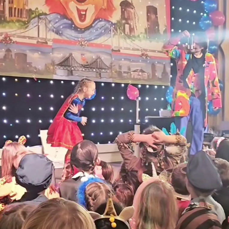 Kinderzauberer Maik auf einer Karnevalsveranstaltung Kind im Kostüm steht auf der Karnevalsbühne, während der Kinderzauberer Maik mit buntem Kostüm mit dem Kind interagiert.