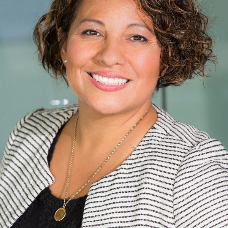 Kindergarten Leitung für Kinderzaubershow Frau mit lockigem Haar, lächelnd, in gestreiftem Blazer vor hellen Hintergrund.
